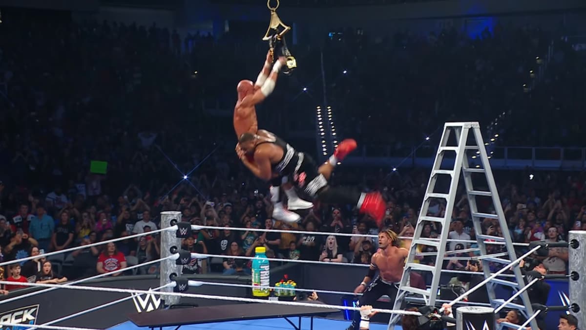 Angelo Dawkins spearing Tommaso Ciampa in mid-air after jumping from a ladder, with a table underneath them