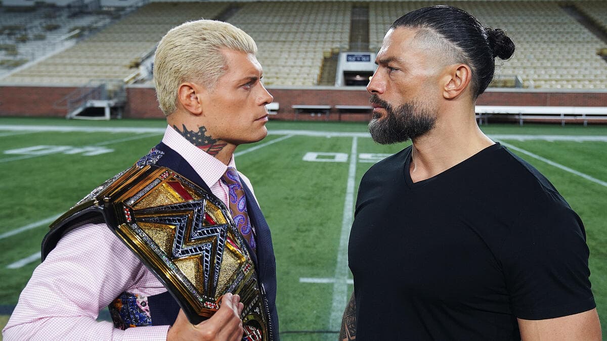 Cody Rhodes (holding the WWE Championship title belt) and Roman Reigns stand on a football field and have a staredown