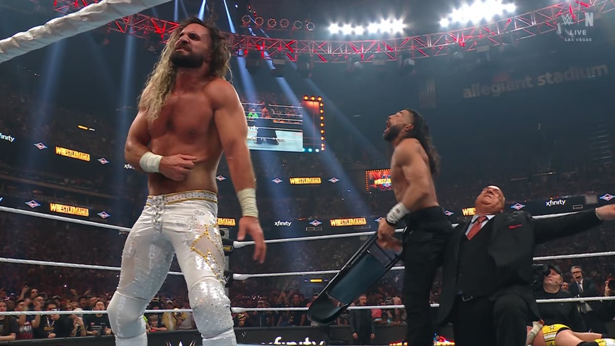 Seth Rollins holds his ribs, as behind him Paul Heyman punches Roman Reigns in the groin in a WWE wrestling ring.