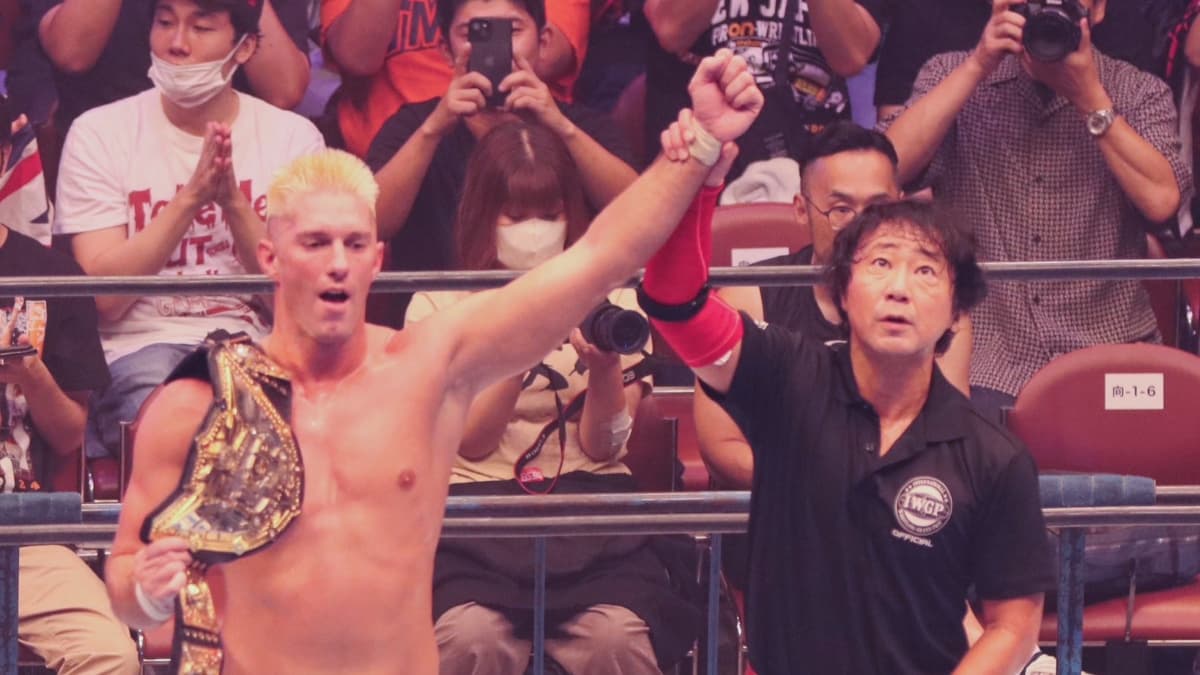 Zack Sabre Jr. with his arm raised by Red Shoes Uno with the IWGP World Heavyweight Title belt across his right shoulder